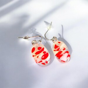 May include: A pair of oval-shaped earrings with a clear base, featuring red and white abstract designs. The earrings have silver-colored hooks and posts. The design is a mix of red and white, creating a unique and artistic look.