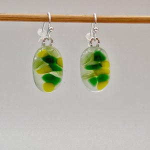 May include: A pair of oval-shaped earrings with silver hooks. The earrings are clear with green and yellow abstract designs inside. The earrings are hanging on a wooden dowel against a white background.