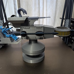May include: A gray 3D-printed model of a vehicle with a rotating base, a gun, and a seat with a blue action figure. The model has a snake-like figure on top and a circular structure on the side.