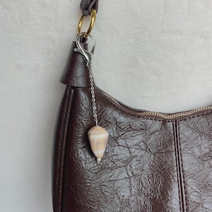 May include: A brown, crescent-shaped handbag with a textured finish. A small, cone-shaped seashell charm hangs from a silver chain attached to the bag's strap. The strap is dark brown with gold-colored hardware.