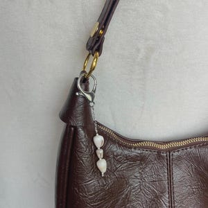 May include: A brown leather shoulder bag with a textured finish, featuring a gold zipper and hardware. A decorative charm with three white, heart-shaped beads hangs from the bag's silver clasp. The strap is dark brown with gold studs.