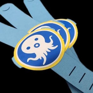 Puede incluir: Pulseras azul claro con tres parches circulares. Cada parche presenta un diseño de pulpo blanco sobre fondo azul, enmarcado por un borde amarillo. Las pulseras tienen una superficie texturizada y líneas negras.