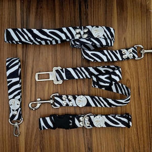 Puede incluir: Cuatro correas para perros de estampado de cebra en blanco y negro con herrajes plateados. Las correas tienen diferentes longitudes y estilos, incluyendo una correa corta con un clip, una correa más larga con una hebilla y una correa con un anillo de metal.