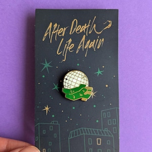 May include: A metal enamel pin featuring a white disco ball held by green hands, attached to a black backing card with the gold text After Death Life Again and a graphic of a city skyline with stars against a dark background.
