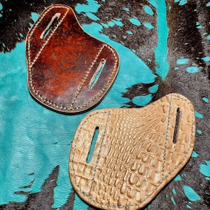 Puede incluir: Dos fundas de cuchillo de cuero, una marrón y otra con estampado de cocodrilo, sobre un fondo de piel de vaca turquesa y marrón.