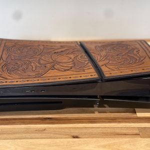 Puede incluir: Una consola de juegos negra con una cubierta de cuero marrón que presenta un intrincado diseño floral. La cubierta está hecha de dos piezas que encajan entre sí.