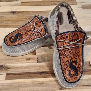 Op de afbeelding: Een paar beige canvas slip-on schoenen met bruine lederen bloemenborduurwerk en de letter "S" op elke schoen.