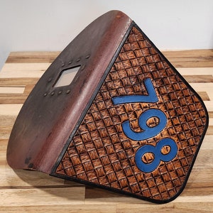 May include: A brown leather plaque with a woven pattern and the number "987" in blue paint.