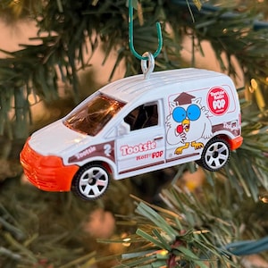 May include: A white and orange Tootsie Roll Pop van ornament with the text "Tootsie Roll Pop" and a cartoon chicken. The ornament is hanging on a Christmas tree with green needles and small white lights.