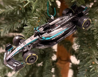 May include: A miniature Mercedes-AMG Petronas Formula One race car ornament in black and silver with teal accents. The car features sponsor logos including Petronas, TeamViewer, and IWC, and is suspended by a green wire hook.