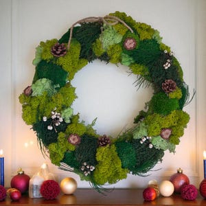 May include: A round wreath composed of various shades of green moss, accented with small pinecones, dried flowers, and white berries. The wreath is suspended by a natural twine loop, suitable for seasonal home decor. The wreath is displayed on a white wall.