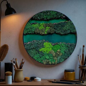 May include: A round wall art piece featuring a layered design of green moss and a teal-colored resin river. The artwork is encased in a black frame and mounted on a light gray wall. The piece is illuminated by a black desk lamp.