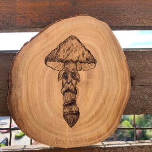 Puede incluir: Un diseño de hongo pirograbado sobre una rodaja de madera circular. El hongo tiene un sombrero detallado y una cara con una larga barba. La veta de la madera es visible y la pieza está colocada sobre un fondo exterior borroso.