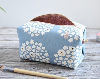 May include: A light blue fabric pouch with a repeating pattern of white fluffy clouds and small cartoon dogs. The pouch features a gold-toned metal zipper closure and a brown interior lining with a white polka dot pattern.