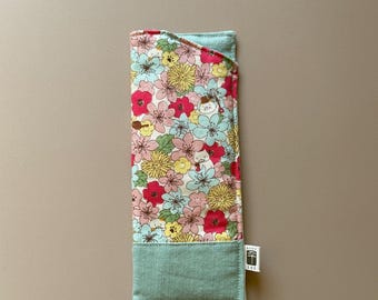 May include: A floral eyeglasses case with a colorful pattern of pink, yellow, and blue flowers. The case has a light blue border and a small tag on the side. The case is rectangular and appears to be made of fabric.