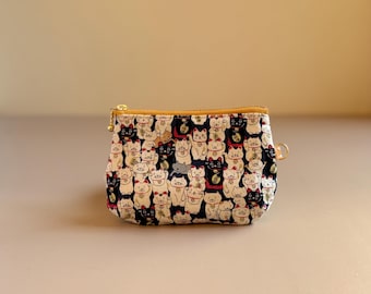 May include: A small, rectangular pouch with a navy blue background covered in a repeating pattern of lucky cat illustrations. The pouch has a gold zipper and a small loop on the side.