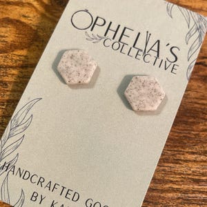 Może przedstawiać: Dwa małe, sześciokątne, nakrapiane, jasnobrązowe kolczyki na kartce z napisem "Ophelia's Collective" i "Handcrafted Goods by Kara".