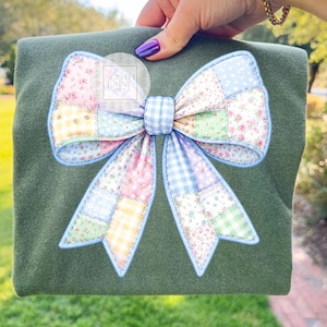 May include: An olive green sweatshirt with a large, colorful bow applique. The bow features a patchwork design with floral, gingham, and solid fabric pieces in pastel colors. The bow is outlined in light blue.