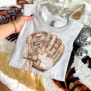 Può includere: Una felpa grigio chiaro con un design a guanto da baseball. Il guanto è un patchwork di tessuti marroni, rosa e beige, con un fiocco al polso. Il design è centrato sulla parte anteriore della felpa.