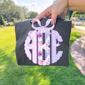 May include: A dark gray t-shirt featuring a monogram design. The monogram is a light purple scalloped circle with the letters ABC in a floral and gingham pattern, accented with a light purple bow.