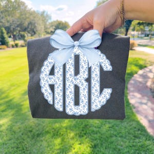 May include: A dark gray shirt with a light blue floral monogram and a matching bow. The monogram features the letters "ABC" in a decorative font, surrounded by a scalloped edge.