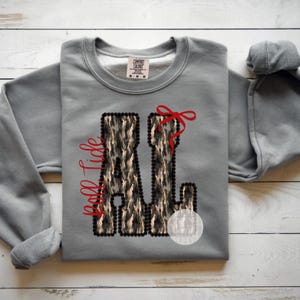 May include: Gray sweatshirt with the letters "AL" in a camouflage pattern, outlined in black, and a red bow. The words "Roll Tide" are written in red script. The sweatshirt is a Comfort Colors brand.