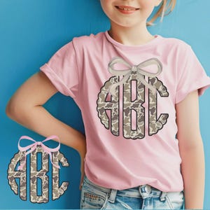 May include: A light pink t-shirt featuring a circular monogram design with the letters "ABC" in a camouflage pattern. A light gray bow is above the monogram. The shirt is a casual style.