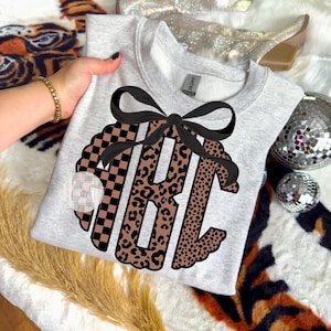 Könnte beinhalten: Hellgraues Sweatshirt mit schwarzer Schleife und Monogramm-Design. Das Monogramm enthält die Buchstaben "ABC" in einer Mischung aus karierten und Leopardenmustern. Das Sweatshirt ist ein Rundhals-Stil.