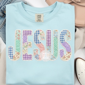 May include: Light blue t-shirt with the word "JESUS" appliqued in a patchwork design. The letters feature various floral and gingham patterns in pastel colors, outlined with a white stitch. The shirt has a crew neck and a Comfort Colors tag.