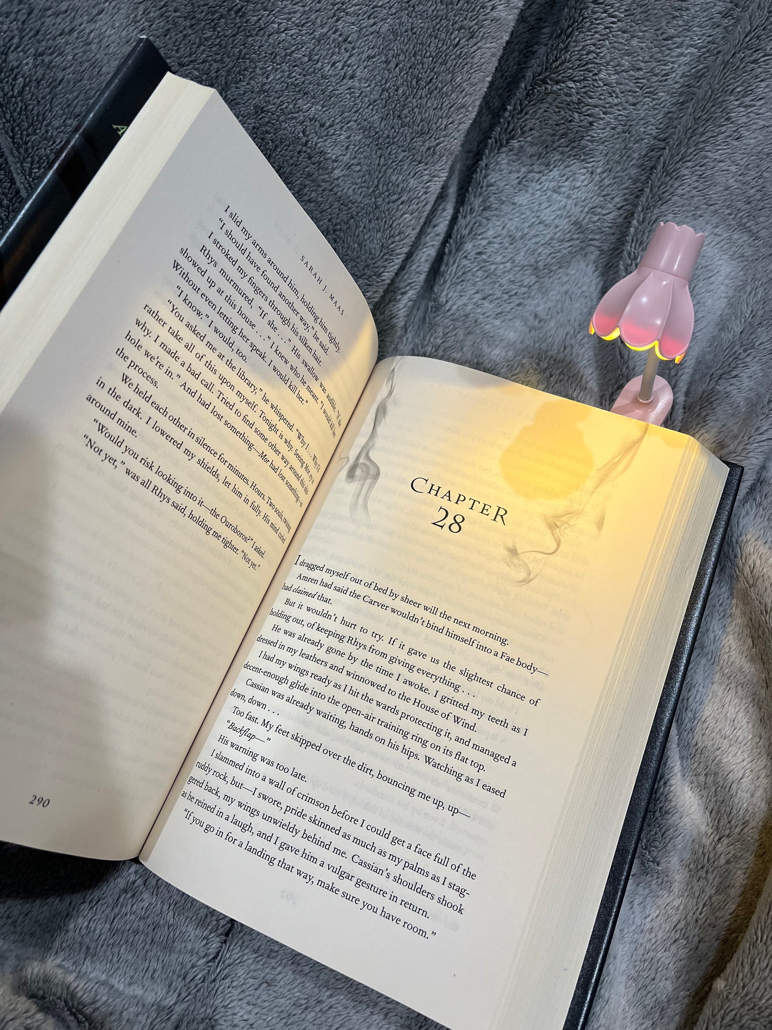 An open book with a small flower shaped booklight attached to the top. It's sitting on a gray blanket.