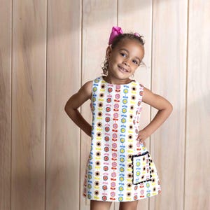 May include: A sleeveless white dress with a colorful sunflower pattern. The dress has a small pocket with a black trim. The girl is wearing pink sandals and a pink bow in her hair. The background is a light brown wooden wall.