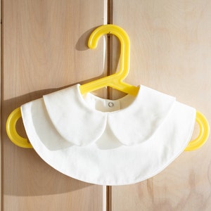 May include: A white cotton peter pan collar with a yellow plastic hanger. The collar is a simple design with rounded edges.