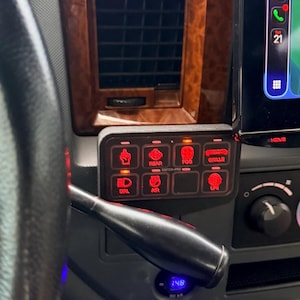 May include: Interior view of a vehicle's dashboard, featuring a steering wheel, navigation screen, and a panel of illuminated switches. The dashboard has a wood-grain finish. A key ring hangs from the steering column. The navigation screen displays a map.
