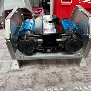 Puede incluir: Un compresor de aire ARB montado en un soporte metálico plateado personalizado. El dispositivo cuenta con dos carcasas de motor cilíndricas azules y negras, un bloque de control central negro con la marca ARB y dos tapas negras circulares.