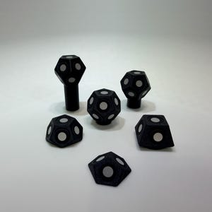 May include: A selection of black, geometric objects with white circular accents. The objects vary in shape, including a tall, cylindrical piece, and several multi-faceted forms. The items are arranged on a white surface.