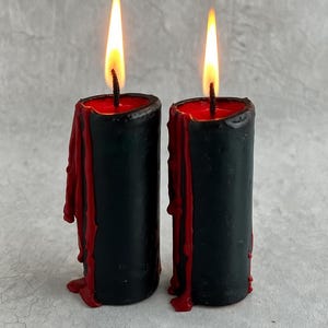 May include: Two lit black candles with red wax dripping down the sides. The flames are bright yellow and orange. The candles have a cylindrical shape and are set against a neutral background.