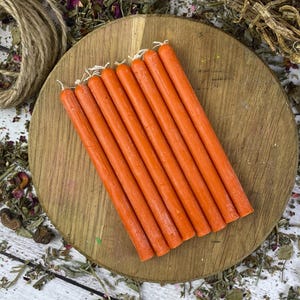 Può includere: Una collezione di candele affusolate arancioni disposte su un tagliere di legno. Sono presenti anche un barattolo di vetro con coperchio di sughero, spago ed erbe essiccate. Lo sfondo è una superficie di legno bianco cosparsa di erbe essiccate e petali di rosa.