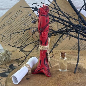 Könnte beinhalten: Rote Kerze mit einem sich umarmenden Paar, einem zusammengerollten Papierstück, das mit Schnur gebunden ist, und einer kleinen Glasflasche mit einem Korkstopfen.