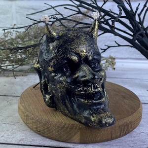 Könnte beinhalten: Schwarze und goldene Kerze in Form eines Teufelskopfes mit Hörnern. Die Kerze steht auf einem Holzsockel.