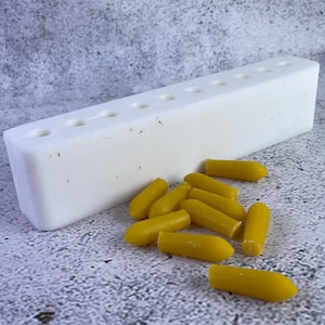 Może przedstawiać: Biała plastikowa forma z 10 okrągłymi otworami, używana do robienia świec. Wokół formy rozrzucone są 12 żółtych woskowych świeczników.