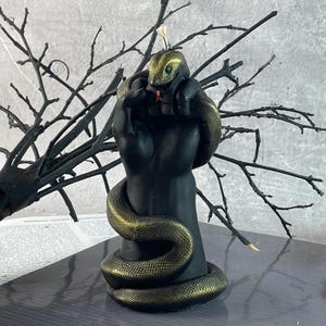 Puede incluir: Una vela de mano negra con una serpiente dorada envuelta alrededor. La cabeza de la serpiente está en la parte superior de la mano y su cola está envuelta alrededor de la muñeca. La vela está sobre una superficie negra.
