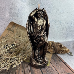 Può includere: Una scultura di candela di colore bronzo che rappresenta una figura alata con un'espressione di tristezza. La figura è seduta con la testa tra le mani e le ali spiegate dietro di lei. La candela è su una superficie di legno.
