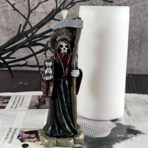 Peut inclure: Figurine de la Faucheuse noire et rouge tenant un sablier, avec une bougie blanche en arrière-plan. La figurine a un visage de crâne et porte une robe noire. La base est dorée. Le fond comprend un journal et un mur gris foncé.