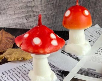May include: Two red and white mushroom shaped candles with white stems. The candles are sitting on a newspaper and autumn leaves.