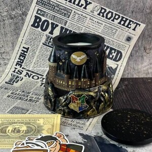 May include: A black candle holder with a Hogwarts castle design, a golden owl emblem, and the Hogwarts crest. The holder sits on a newspaper with the headline "Daily Prophet". Includes stickers and a black lid.