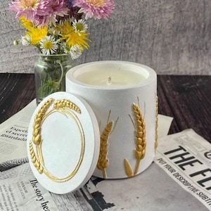 Puede incluir: Una vela blanca en un recipiente cilíndrico con tapa, decorada con espigas de trigo doradas. La vela está sobre un periódico con un pequeño ramo de flores en un tarro de cristal.