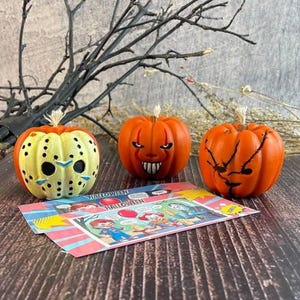 Può includere: Tre zucche decorative di Halloween con facce intagliate. Una è gialla con un design a maschera da hockey, le altre sono arancioni. Una colorata carta a tema Halloween è di fronte alle zucche.