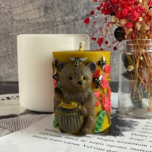 May include: A yellow candle with a brown bear holding a honey pot. The candle is decorated with bees and flowers. A white candle and a glass jar with red flowers are in the background.