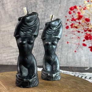 Two black handcrafted female body candles styled like classical sculptures, placed on a round wooden platform next to a glass vase with red and white roses