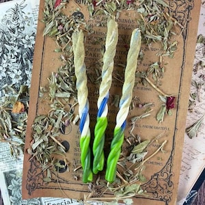 Puede incluir: Tres velas cónicas trenzadas con un patrón de degradado en verde claro, verde oscuro, azul y blanco. Las velas están dispuestas verticalmente sobre un papel de estilo vintage impreso con texto y rodeadas de hierbas secas esparcidas.